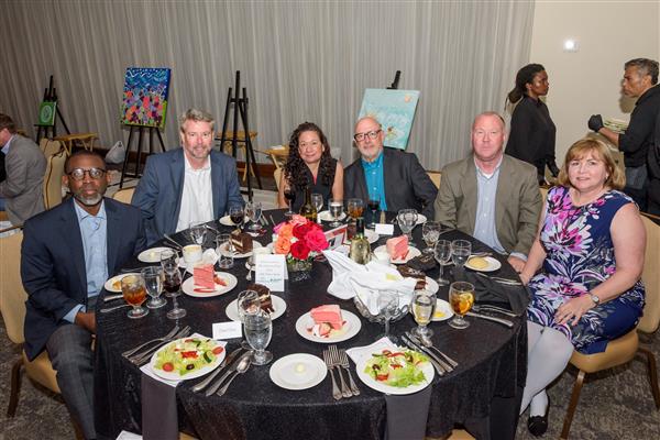 A group of six well-dressed adults pose for a photo at a formal dinner event. They are all seated at the table. The table is set with glasses, plates of cake, salads, rolls, and bottles of wine and water. The background shows a large room filled with other guests seated at tables, with artwork on ea